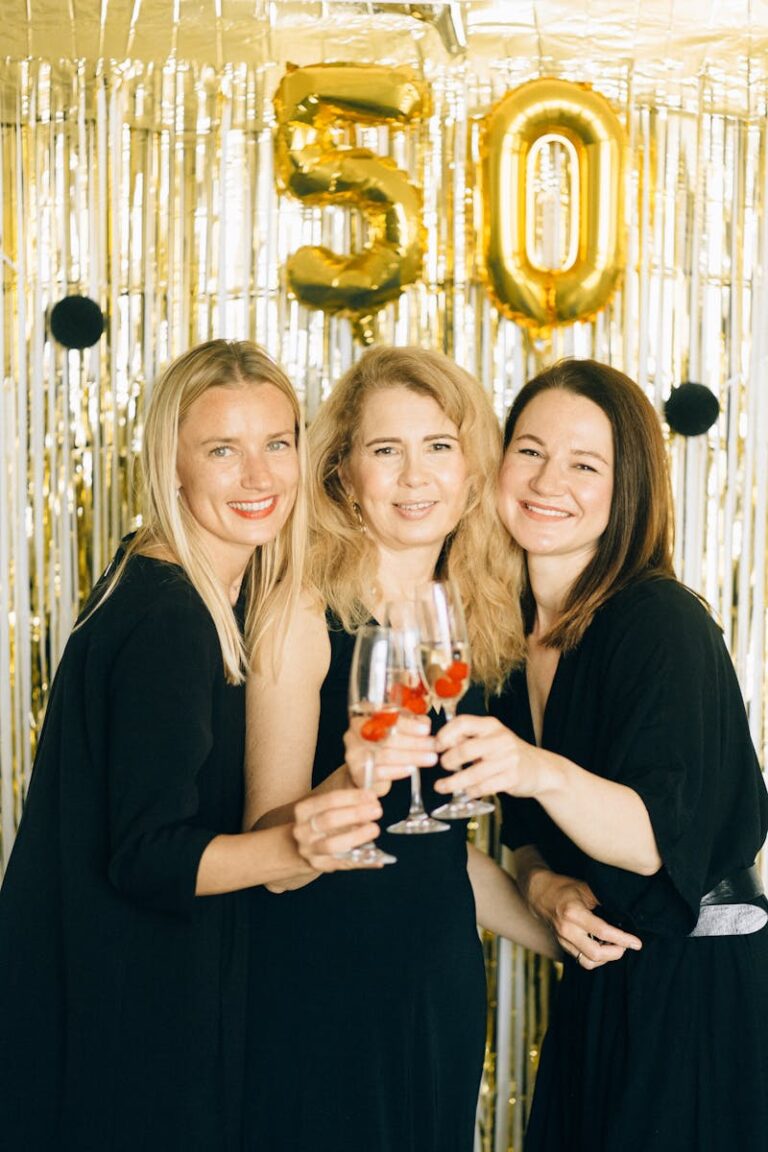 Three women celebrating a 50th birthday with champagne, wearing black dresses and smiling.